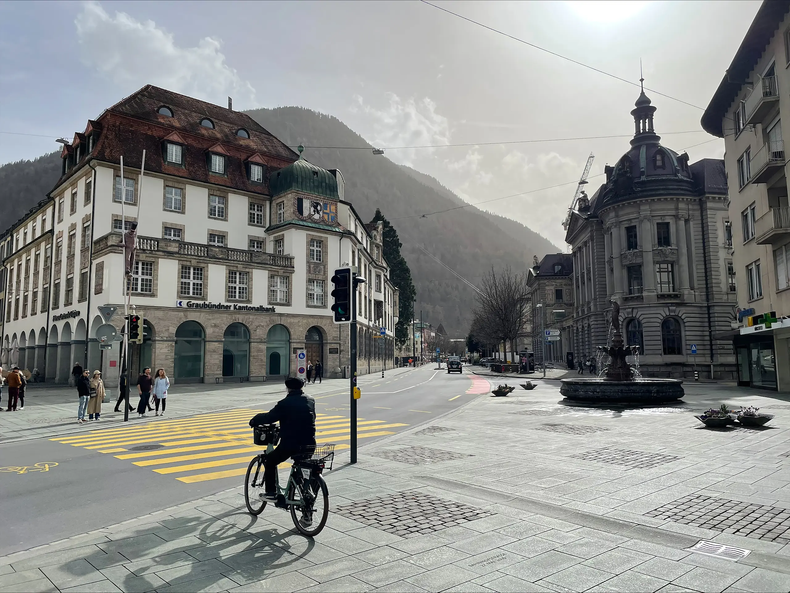 Der Postplatz in Chur, Schweiz, im Jahr 2025. Eine urbane Umgebung mit klassischen Gebäuden, Autoverkehr und wenigen Grünflächen.